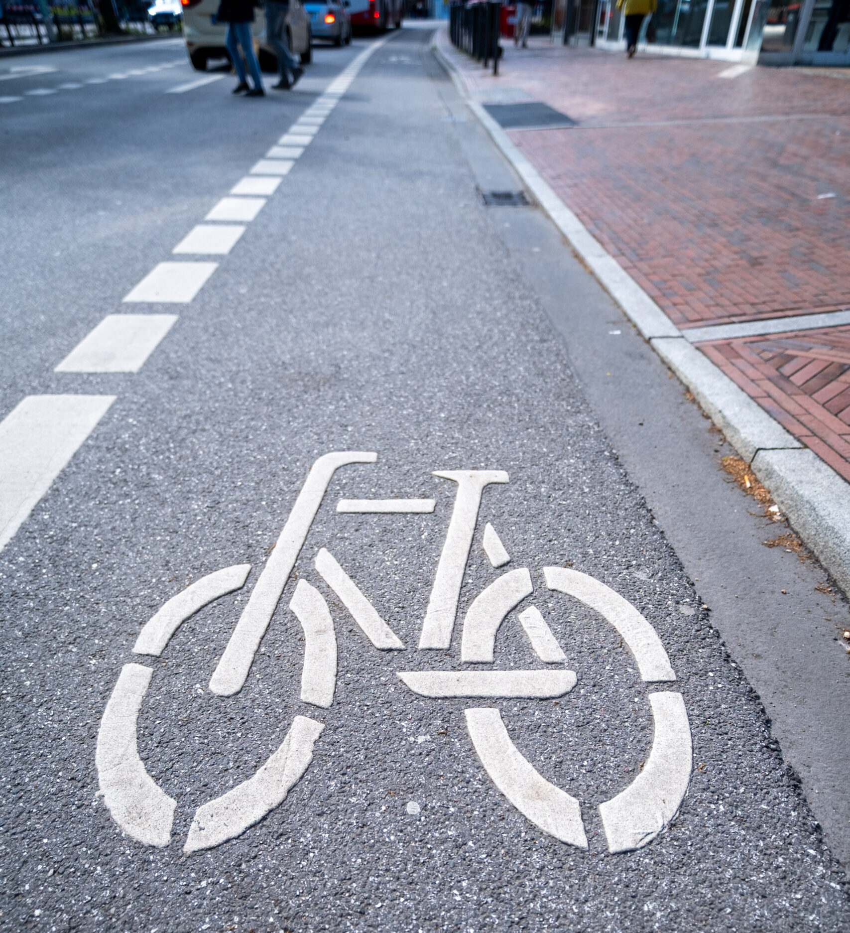 Ein Fahrradsymbol auf einer Straße, im Hintergrund sind Autos und Menschen zu erkennen. Das Symbol markiert einen Fahrradschutzstreifen mit gestrichelter Linie als Grenze zur Autofahrbahn.